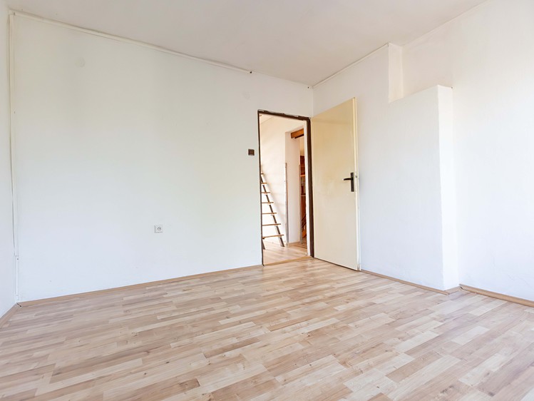 Prázdný pokoj s dřevěnou podlahou, bílými stěnami a otevřenými dveřmi.