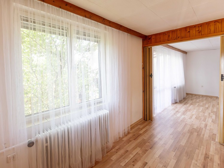 Prosvětlený pokoj s dřevěnou podlahou, radiátorem a průchodem do další místnosti.