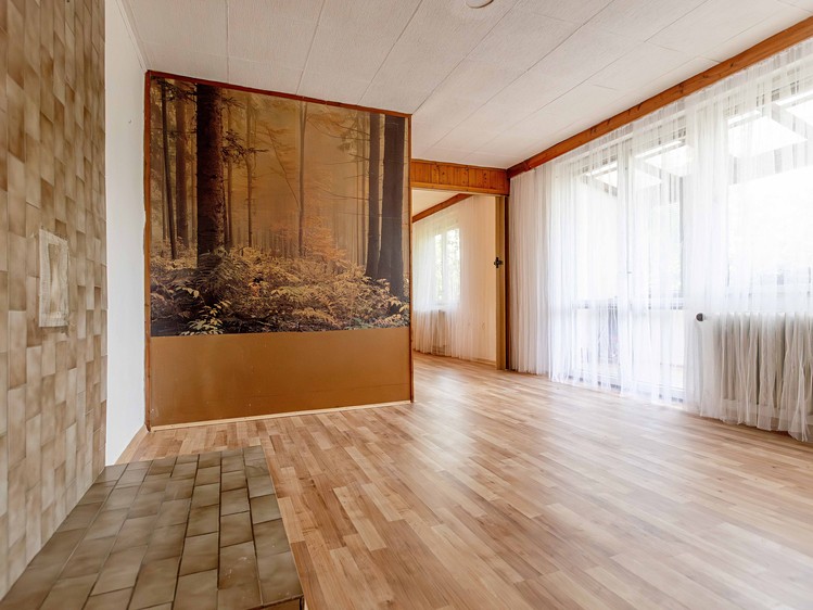 Interiér s dřevěnou podlahou, fototapetou lesa a velkým proskleným oknem se záclonami.