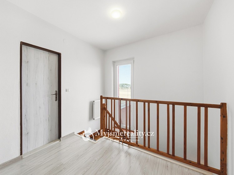 Podesta se světlou podlahou, dřevěným zábradlím, dveřmi a vstupem na balkon.